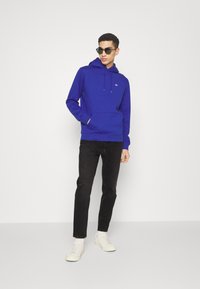 Blauwe hoodie met een voorzak in kangoeroestijl, gecombineerd met zwarte jeans en witte sneakers. Model draagt een zonnebril.