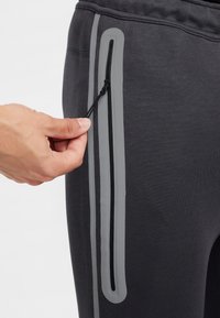 Pelēkas sporta bikses ar vertikālu rāvējslēdzēja kabatu, kurā ir melns vilkšanas aukla un atstarojoši pelēki elementi sānu šuvē.