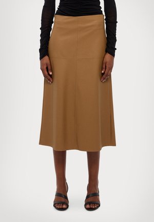 Femme portant une jupe midi marron évasée, associée à des sandales noires à talons et un haut noir froncé à manches longues, debout devant un fond blanc.