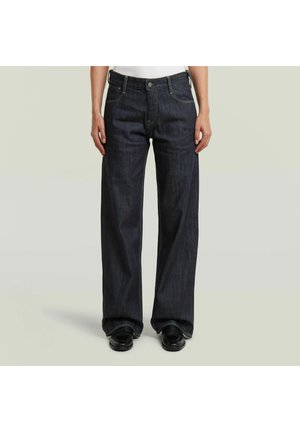 Persoon die donkere wijde jeans en zwarte schoenen draagt, staand tegen een effen lichte achtergrond.