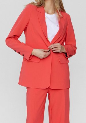 Femme boutonnant un blazer corail vif porté avec un pantalon assorti et un t-shirt blanc à motif floral sur un fond uni.