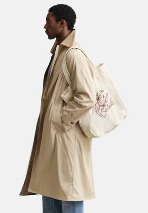 Mann trägt beige Trenchcoat und trägt weiße Canvas-Tragetasche mit rotem Hummer und dem Text "Seaside" über der Schulter.
