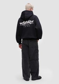 Černá mikina s kapucí a bílým grafickým nápisem na zádech, kombinovaná s tmavými cargo kalhotami. Měkká tkanina, volný střih a žebrované manžety.