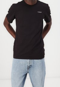 Zwart T-shirt met korte mouwen, gemaakt van katoen, met een klein wit logo op de borst en een klassieke ronde hals. Draagt men met lichtblauwe jeans.