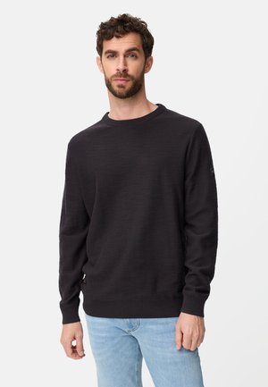 Mężczyzna z kręconymi włosami i brodą, ubrany w czarny sweter z długim rękawem i okrągłym dekoltem oraz jasnoniebieskie dżinsy, stojący na tle jednolitego tła.