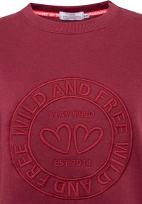 Bordowy sweter z okrągłym dekoltem, z wypukłym haftem tekstu "STAY WILD" oraz okrągłym wzorem z sercami.