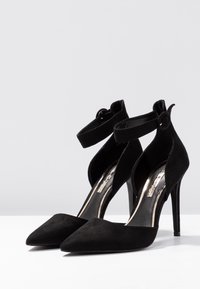 Černé semišové lodičky na vysokém podpatku s špičatou špičkou a kotníkovými pásky. Mají výřezový design a stříbrné detaily na stélce.