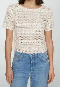 Luźna, kremowa bluzka o kroju cropped z teksturowanym, otwartym wzorem szydełkowym i falbaniastymi brzegami, połączona z jasnoniebieskimi dżinsami.