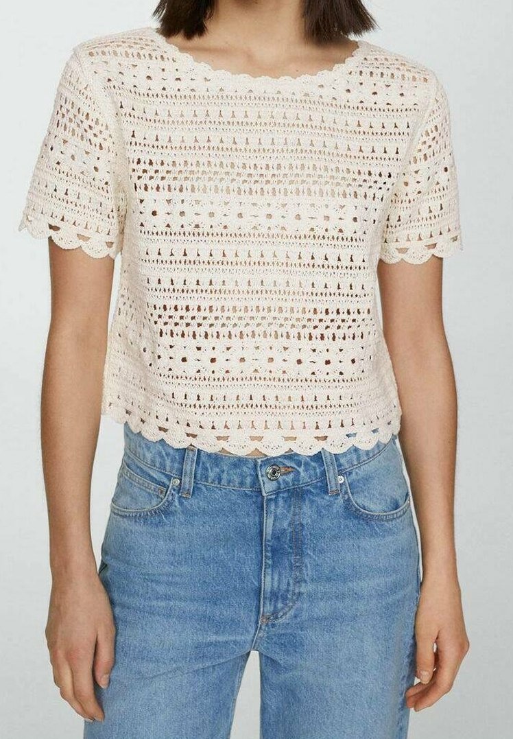 Luźna, kremowa bluzka o kroju cropped z teksturowanym, otwartym wzorem szydełkowym i falbaniastymi brzegami, połączona z jasnoniebieskimi dżinsami.