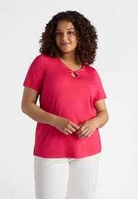 T-shirt rosa a maniche corte in materiale morbido con scollatura a chiave decorata da cuciture decorative. Abbinato a pantaloni bianchi.