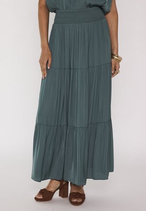 Femme portant une longue jupe bleu sarcelle avec des volants froncés et une taille ajustée, associée à des sandales marron à talons blocs et des bijoux en or.