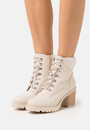 Beige enkellaarsjes met hak, veters aan de voorkant, metalen oogjes, zijrits en omgeslagen faux fur kraag, gedragen op blote benen.