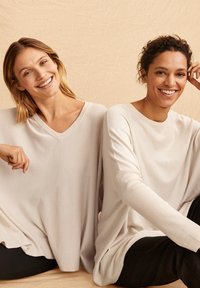 Zwei Frauen tragen lockere, hellbeige Pullover mit V-Ausschnitten. Der Stoff wirkt weich mit einer strukturierten Oberfläche, kombiniert mit dunklen Hosen.