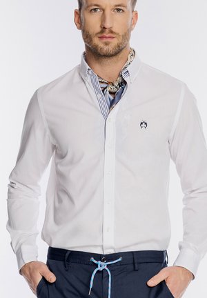 Mężczyzna ubrany w białą koszulę zapinaną na guziki z haftowanym logo, wzorzystą apaszkę na szyi oraz granatowe spodnie ze sznurkiem w kolorze niebieskim, z rękami w kieszeniach.