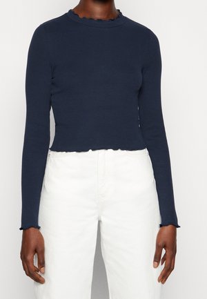 Personne portant un haut côtelé bleu marine ajusté à manches longues avec des détails de bordure en forme de feuille de laitue, associé à un pantalon blanc taille haute.
