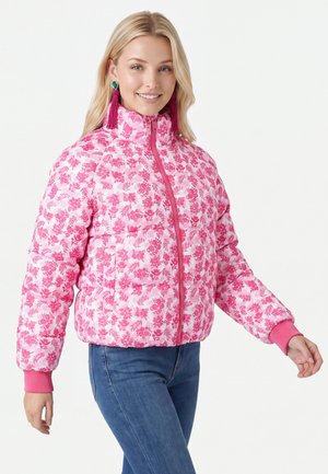 Junge Frau trägt eine rosa gesteppte Jacke mit floralem Muster und hohem Kragen, blaue Jeans und lange Quastenohrringe, lächelt vor weißem Hintergrund.