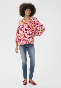 Gėlių bliuska raudonos, rožinės ir oranžinės spalvų su mandarinų apykakle ir pūstomis rankovėmis, sujungta su šviesiai mėlynomis siaurėjančiomis džinsomis ir raudonomis balerinomis.