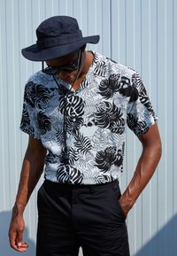 Short-sleeve shirt with a black and white leaf pattern, featuring a subtle skull design, paired with black trousers and a navy bucket hat.