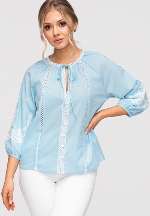 Femme portant un chemisier bleu clair avec une bordure en dentelle blanche, des boutons sur le devant, un col à nouer et des manches 3/4 blanches brodées, associées à un pantalon blanc.