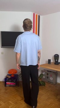 Vaaleansiniset ylisuurikokoiset T-paita, mustat leveälahkeiset housut, näkyvät tatuoinnit, moderni sisustus televisiolla, oranssilla lampulla ja koristekasvilla.