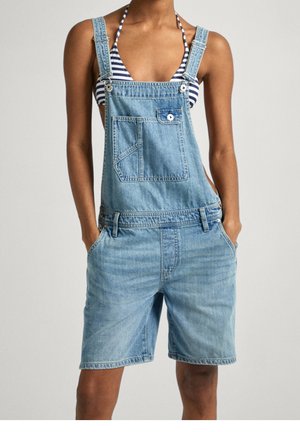 Persoon draagt lichtblauwe denim korte tuinbroek met voorvak en gestreept bikinibovenstuk eronder, handen in de zakken.