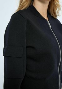 Donna che indossa una giacca di maglina nera con una cerniera argentata frontale e un pattina sulla tasca della manica superiore, su uno sfondo chiaro e semplice.