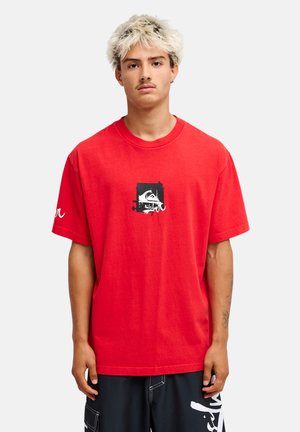 Młody mężczyzna o jasnych, kręconych włosach w czerwonym t-shircie z nadrukiem i czarnych szortach stoi na tle jednolitego jasnego tła.