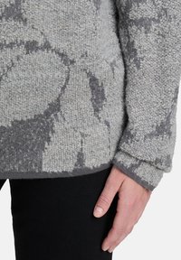 Grauer Strickpullover mit einem strukturierten Blumenmuster, gerippten Bündchen und einem lässigen Schnitt. Das Material wirkt weich und warm.