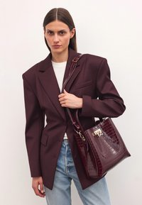 Burgundy läderhandväska med krokodilstruktur, med gyllene detaljer och lång rem. Modellen bär en mörk kavaj och ljusa jeans.
