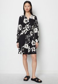 Schwarzes Blumen-Wickelkleid mit langen Ärmeln, verziert mit großen weißen Blumen und einem taillierten Schnitt. Tragen mit schwarzen flachen Sandalen.