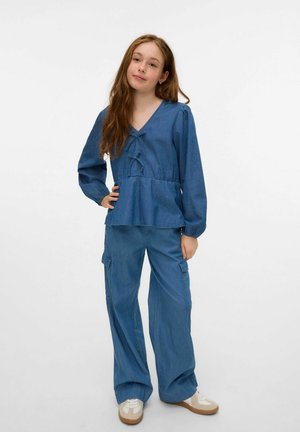 Denim top med v-udskæring, lange ærmer og peplum-silhuet; parret med vide denimbukser og lyse sneakers.