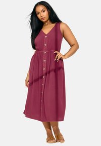 Vestido burdeos sin mangas con escote en V, botones en la parte delantera, cintura ceñida y falda fluida, hecho de una tela suave. Llevado con sandalias color tostado.