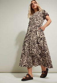 Beige maxi-mekko mustalla abstraktilla pyörteellä, lyhyet hihat ja taskut. Yhdistetty tummanruskeisiin liukuviin sandaaleihin, joissa on niittejä.