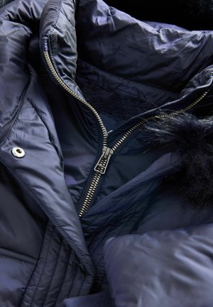 Marineblaue Steppjacke mit silbernem Reißverschluss, Fellbesatz und gestepptem Design. Das Innere hat eine weiche Fütterung und dezente Steppdetails.