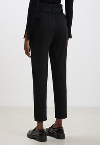 Pantaloni neri con una vita alta e testurizzata, dotati di passanti per cintura. Design a gamba affusolata con lunghezza corta. Indossati con eleganti scarpe nere.