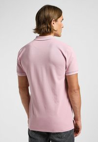 Polo shirt rose avec une texture douce, des manches courtes et un col. Les détails notables incluent des coutures contrastées et des fentes sur les côtés à l'ourlet.