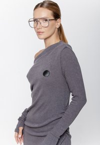 Szary sweter prążkowany z jednym odkrytym ramieniem, długimi rękawami i okrąłym czarnym akcentem z przodu. Gładka tekstura.