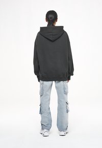 Persona con el cabello recogido, que lleva una sudadera negra de gran tamaño, unos pantalones cargo azul claro y zapatillas blancas, de pie de espaldas sobre un fondo blanco.