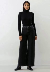 Crna rebrasta turtleneck majica u kombinaciji s širokim crnim trapericama. Oba komada odlikuju se glatkom teksturom i visokim strukom, a upotpunjuju ih sjajne crne cipele.