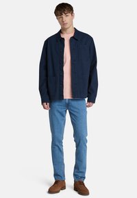 Navyblauwe katoenen jas met knopen, twee voorzakken, gedragen over een lichtroze t-shirt, gecombineerd met blauwe jeans en bruine schoenen.