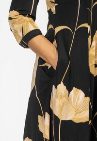 Czarna sukienka z dużymi beżowymi wzorami kwiatowymi, z rękawami trzy czwarte i bocznymi kieszeniami. Gładki materiał o luźnym kroju.