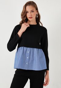 Top nero a maniche lunghe con peplum a righe blu e bianche, caratterizzato da tre bottoni davanti. Texture liscia, blocchi di colore a contrasto.