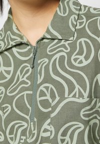 Giacca con cerniera color verde oliva con motivo grafico di forme astratte in verde chiaro. Dettagli distintivi includono un colletto alto e una tasca frontale.