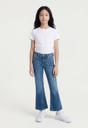 Fille aux longs cheveux noirs portant un t-shirt blanc, un jean évasé bleu et des baskets blanches, debout devant un fond clair uni.