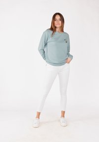 Felpa azzurra con scollo rotondo e ricamo floreale sul lato sinistro del petto, abbinata a jeans skinny bianchi e sneakers bianche.