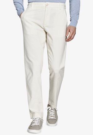 Mężczyzna noszący proste, kremowe spodnie, szare trampki na sznurowadła oraz jasnoniebieską koszulę z długim rękawem, z jedną ręką w kieszeni.