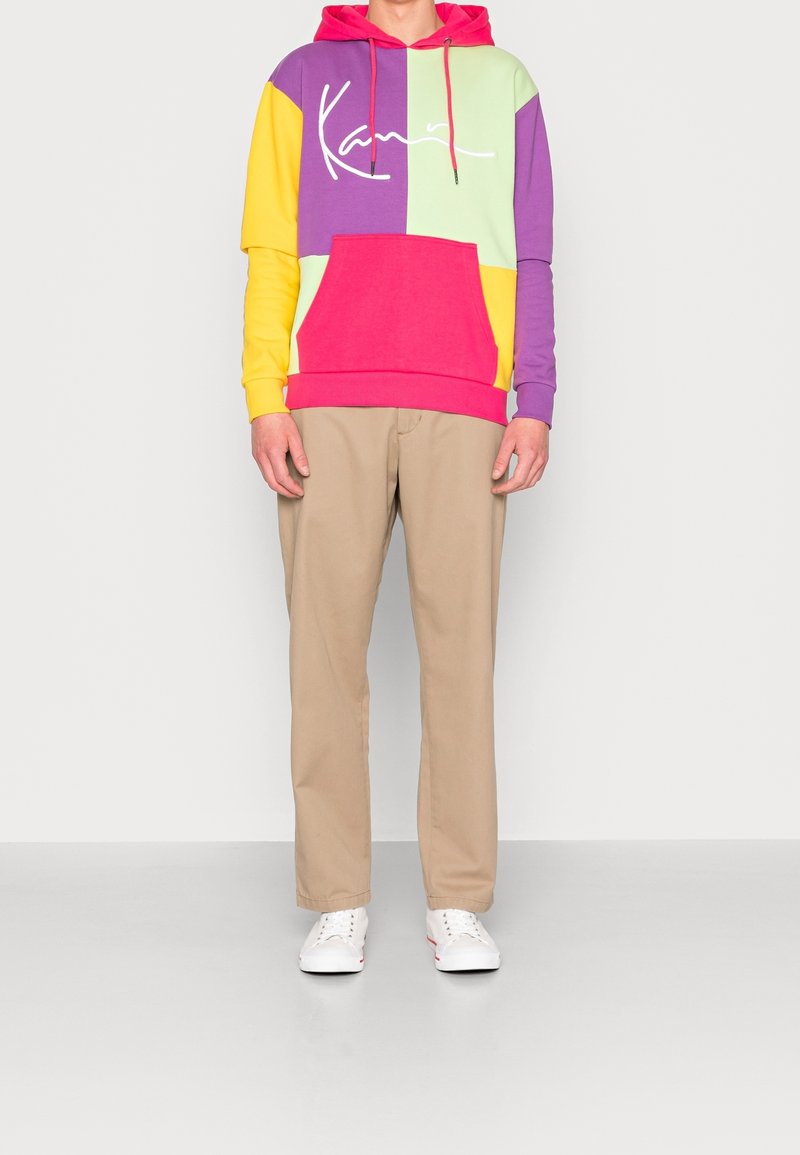 Sudadera con capucha colorida con secciones moradas, verdes, amarillas y rosas; presenta un bolsillo frontal y un logo en texto blanco. Combinada con pantalones beige y zapatillas blancas.