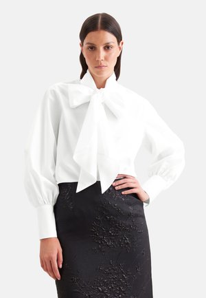 Femme portant une blouse blanche avec un grand nœud et des manches bouffantes, associée à une jupe noire ornée de broderies florales.