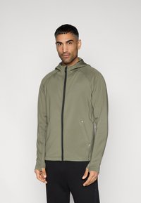 Φούτερ σε ελιά πράσινο με φερμουάρ μπροστά, πλευρικές τσέπες και εφαρμοστή σχεδίαση. Διαθέτει μανίκια ράγκλαν και υφή από ελαφρύ ύφασμα.