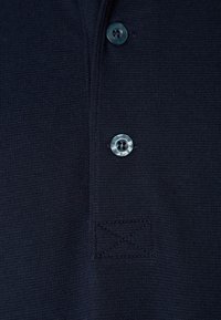 Laivaston sininen poolopaita, jossa on teksturoitu kangas, kolme kontrastin värisellä ompeleella varustettua nappia ja ommeltu yksityiskohta nappilistan lähellä.